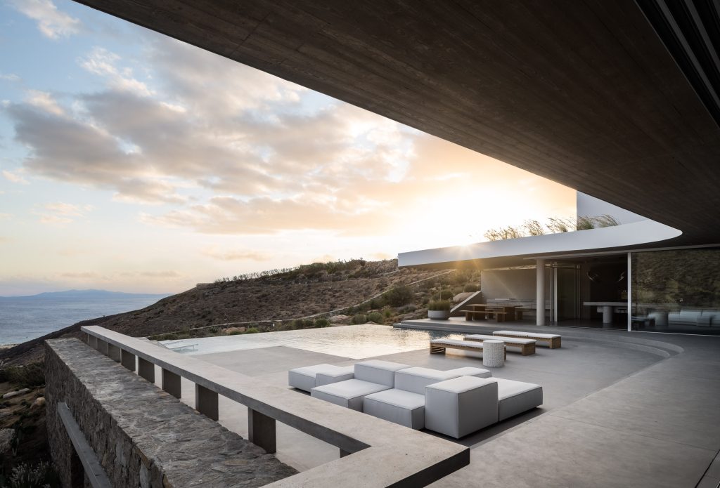 O elemento central da composição é um muro de pedra de 62 metros, construído com granito local, que acompanha as curvas naturais do terreno. A residência encaixa-se de forma orgânica nesse eixo, abrindo-se para sul com vistas amplas sobre o mar, e para este, diante de uma colina rochosa com muros de pedra histórica.
Todas as coberturas da AIMASIA Residence foram transformadas em telhados verdes com vegetação autóctone, promovendo isolamento térmico e integração paisagística. O betão aparente, produzido com cimento de baixo teor de carbono, confere durabilidade e massa térmica à estrutura — armazenando calor no inverno e refrescando os espaços no verão.